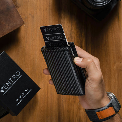 A hand holds a black carbon-fiber-textured Ventro wallet with two black Ventro cards partially pulled out, set on a wooden tabletop with a Canon camera, a brown leather pouch, and a black Ventro box nearby.